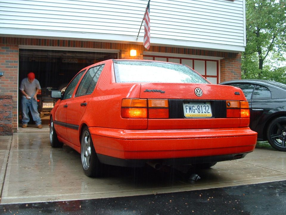 Rear view of an orange 1994 Volkswagen Jetta parked in a driveway, with a black car to the right