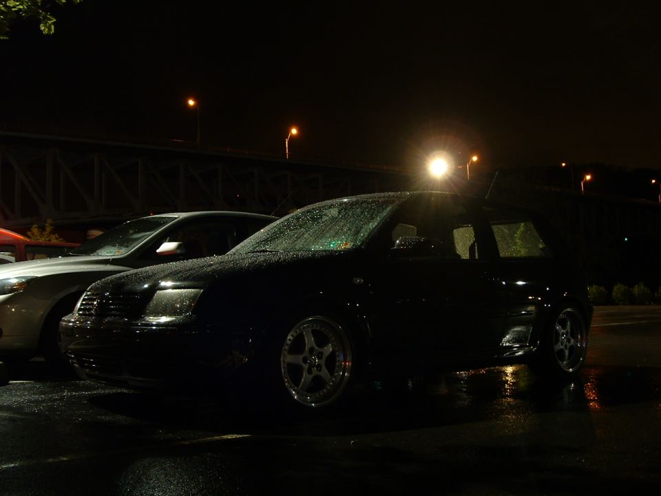 Black sedan parked next to another car at night, rain on the windshield