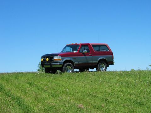 1995 Ford Bronco - A two-tone Ford Bronco sits on a grassy knoll, its red and gray paint contrasting with the bright blue sky.