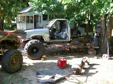 1985 Ford Bronco - A partially disassembled pickup truck sits in a yard with parts and equipment scattered around.
