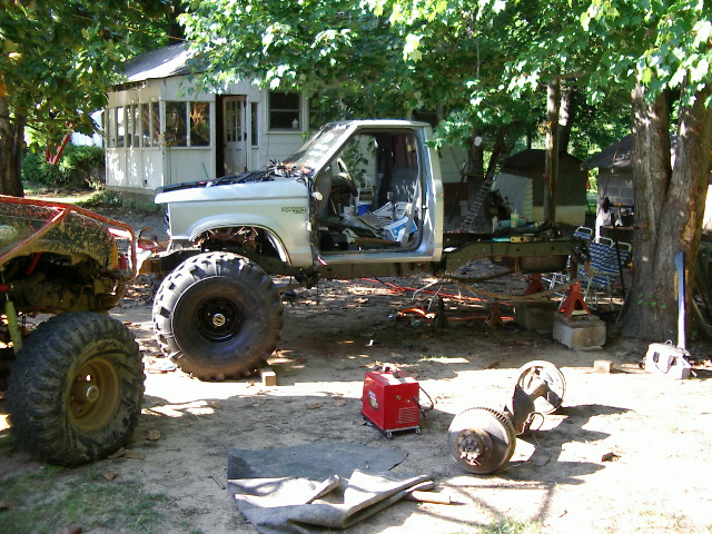Pickup truck chassis with large tires, open doors, and hood. Another off-road vehicle is to the left. Parts and a welder are on the ground