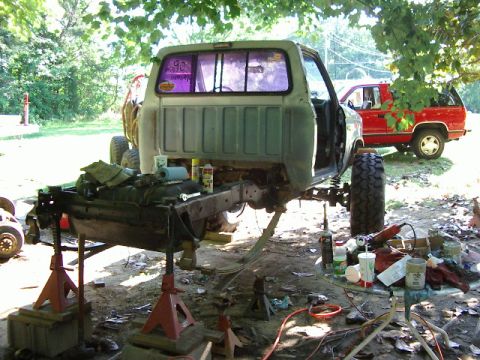 1985 Ford Bronco - A pickup truck body is elevated on jack stands, undergoing repairs or modifications, with another red truck visible in the background.