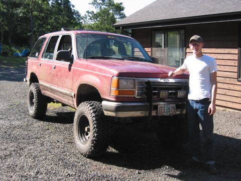 1991 Ford Explorer - A man stands beside a red 1991 Ford Explorer modified with large tires and a lifted suspension.