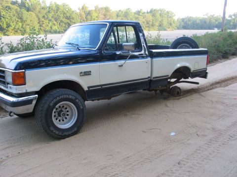 1989 Ford F-150 - A two-tone cream and dark blue pickup truck is parked on a dirt road with its rear passenger-side wheel missing.