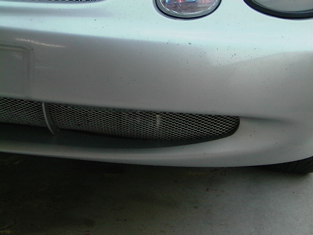 Silver car bumper with a black mesh grille and a portion of a headlight visible