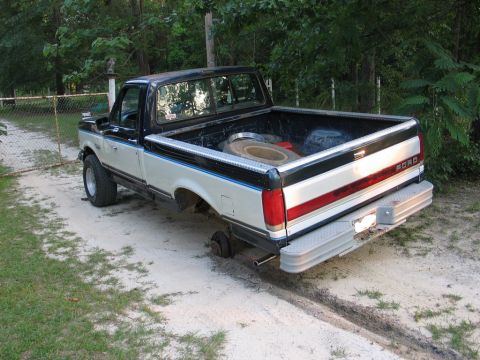1989 Ford F-150 - A two-tone Ford pickup truck is parked on a dirt road with its rear passenger-side wheel missing.