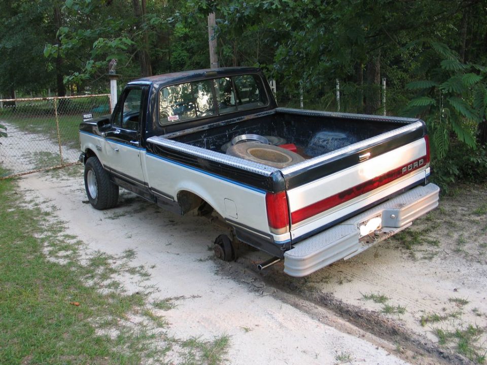 Two-tone Ford pickup truck missing rear passenger-side wheel, with spare tire in bed