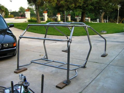 1985 Ford Bronco - A custom metal roll cage sits on a concrete driveway, with a black car parked nearby and welding equipment in the foreground.