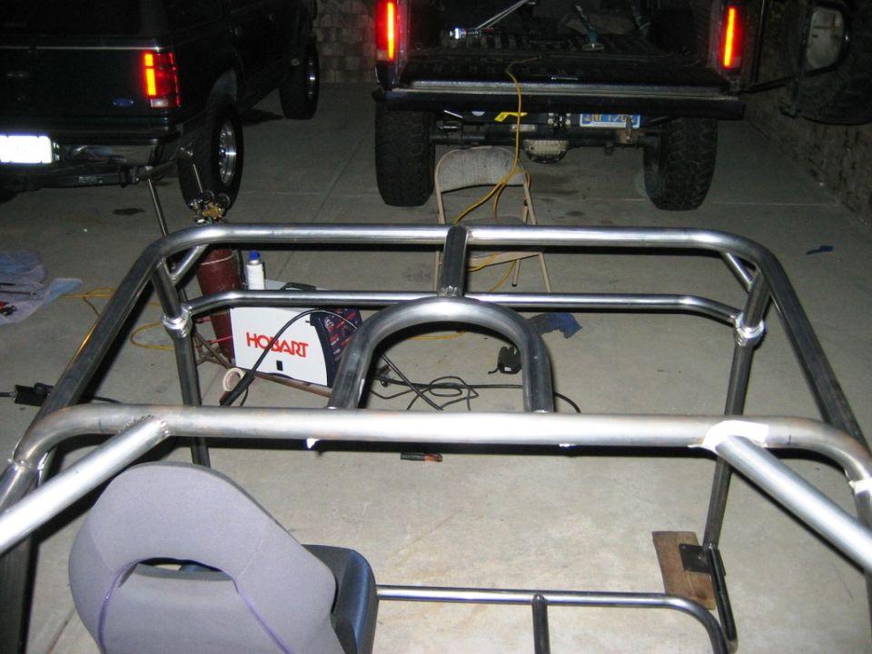 Bare tubular steel roll cage frame under construction in a garage with a Hobart welder and seat.