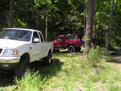 1994 Ford Bronco - Two trucks, a red Bronco and a white F-150, are parked off-road in a forest.