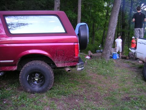 1994 Ford Bronco - A red SUV is parked in a wooded area next to a campfire scene with people gathered.