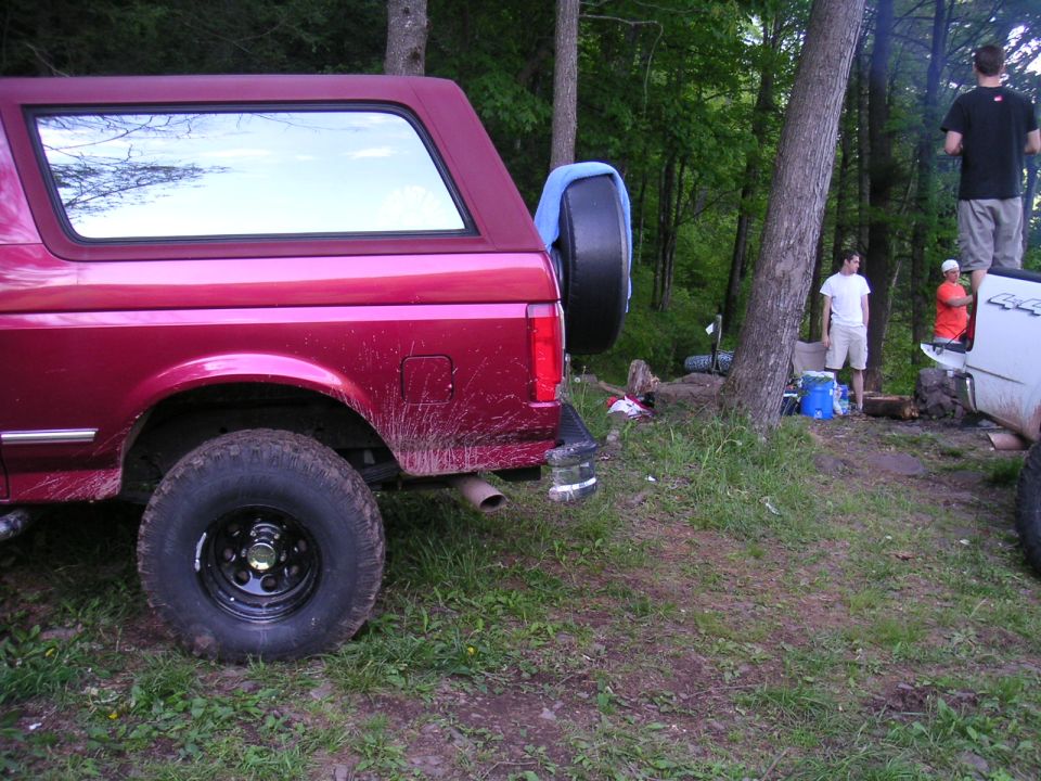 Red SUV with spare tire on back, parked in a wooded area. Another white truck is nearby with people standing around a campfire