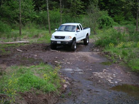 1994 Ford Bronco - White pickup truck navigates a challenging muddy and rocky path through a wooded area.