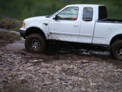 1994 Ford Bronco - A white pickup truck navigates a rocky, muddy terrain near a body of water.