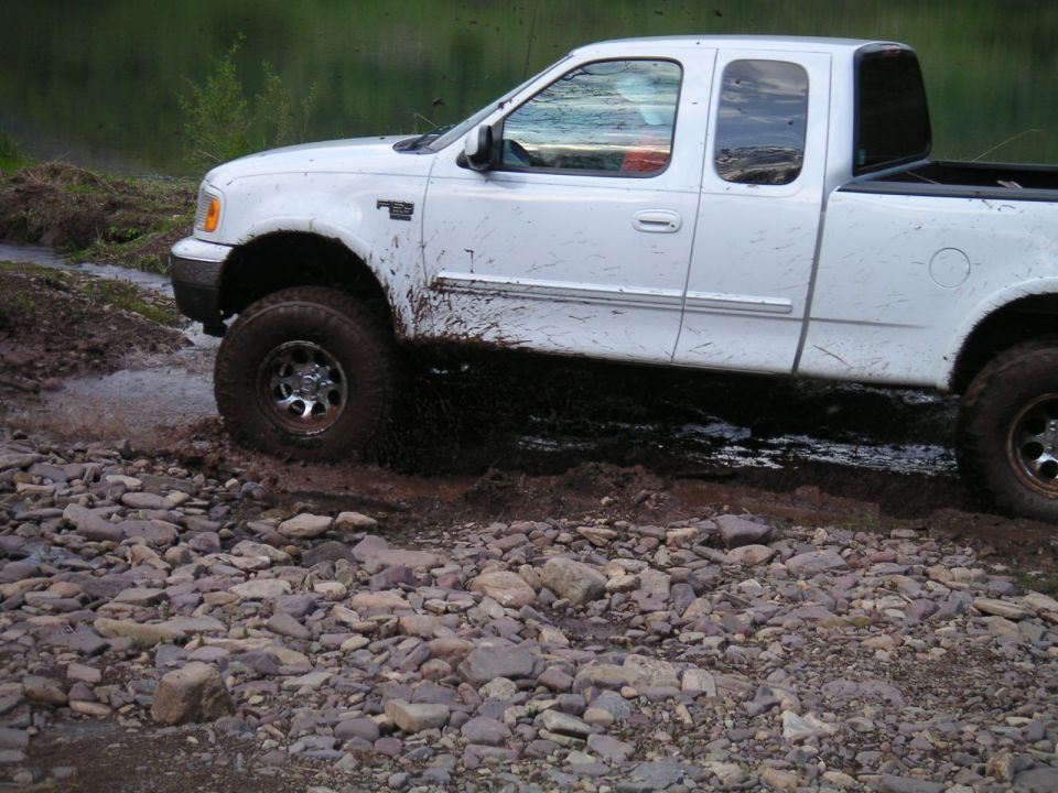 White pickup truck with mud splattered on its side, driving over rocks and through water