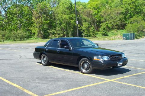 1999 Ford Crown Victoria - A black sedan is parked in an empty lot with trees in the background.