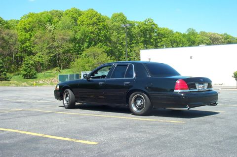 1999 Ford Crown Victoria - This black sedan is parked in an asphalt lot with trees and a building visible in the background.