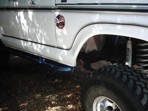 1978 Ford Bronco - Close-up of a white SUV's fender and tire, a patriotic decal and off-road modifications.
