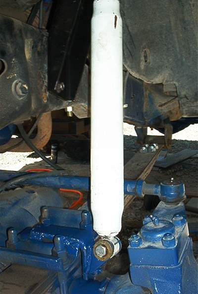 1986 Ford Bronco - Underside view of a vehicle's front suspension showing a white steering damper/stabilizer mounted vertically, connected to blue-painted axl…