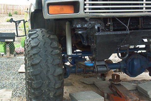 1986 Ford Bronco - Low front-end shot of a lifted truck showing the front axle, leaf spring suspension, steering linkage, and a large mud-terrain tire. An amb…