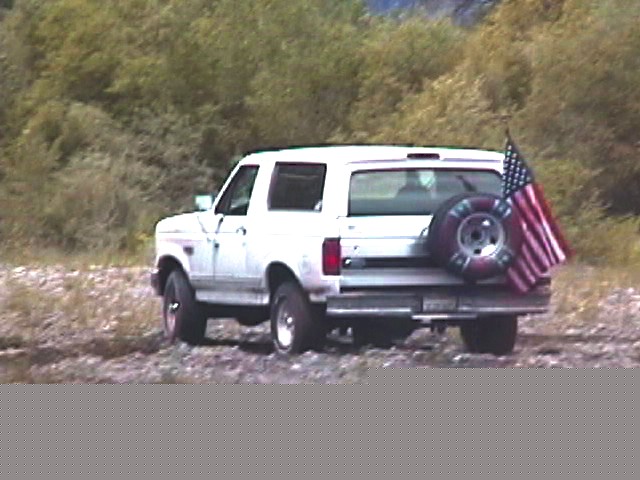 White 1993 Ford Bronco parked on a dirt road with trees in background, American flag on spare tire