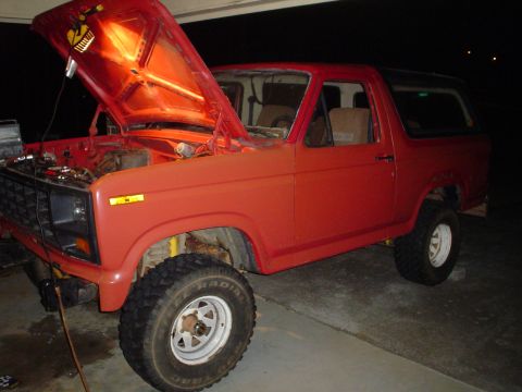 1981 Ford Bronco - The front end of a red SUV is visible with the hood open, revealing the engine bay under a work light.