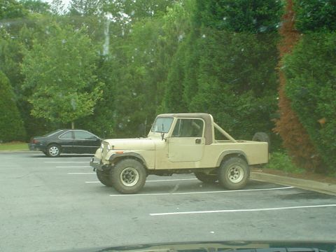 1981 Ford Bronco - A tan Jeep CJ-7 is parked in a lot next to a black sedan, with trees in the background.