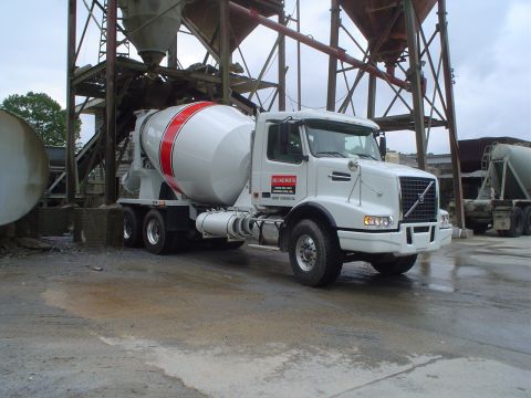 1981 Ford Bronco - A white concrete mixer truck is parked on wet ground next to industrial silos.