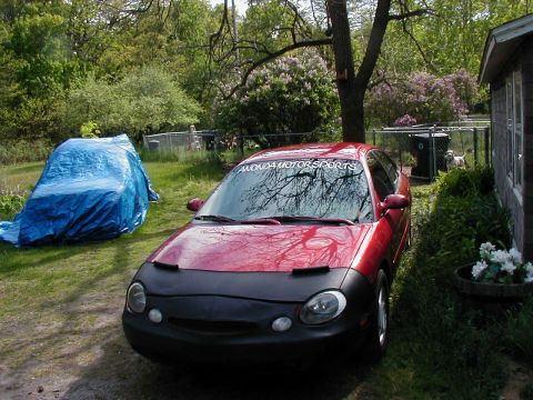 1997 Ford Taurus - A red 1997 Ford Taurus sedan is parked in a yard, partially covered by a black car bra and a windshield banner.