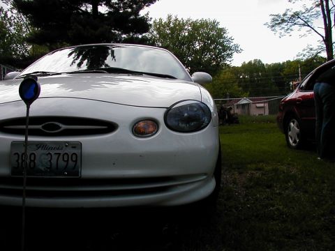 1997 Ford Taurus - Front view of a white 1997 Ford Taurus, showing its distinctive headlights and grille. Another vehicle is visible in the background.
