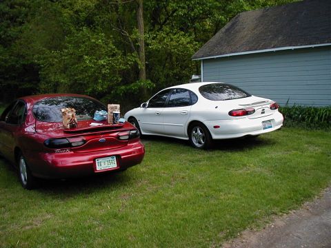 1997 Ford Taurus - Two Ford Taurus sedans, a red one and a white one, are parked on a grassy area near a house.