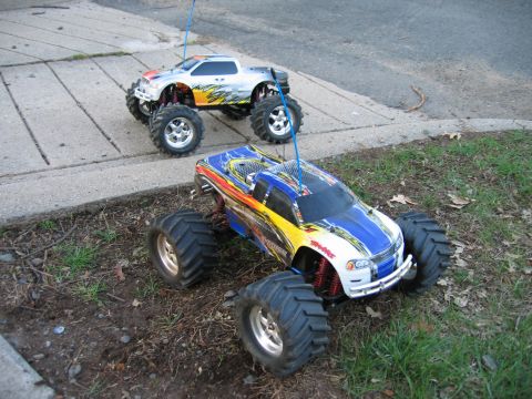 1996 Ford Crown Victoria - Two remote control monster trucks are parked on the grass and concrete, one in the foreground and another behind it.