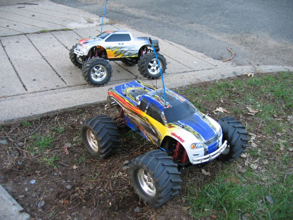 Two RC Traxxas monster trucks with flame graphics on grass and sidewalk