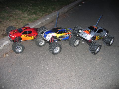 1996 Ford Crown Victoria - Three remote control monster trucks with flame decals are lined up on a paved surface.