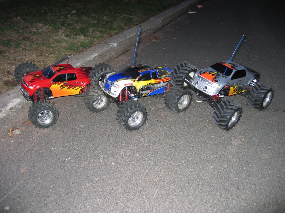 Three RC monster trucks with flame-painted bodies lined up on pavement at night