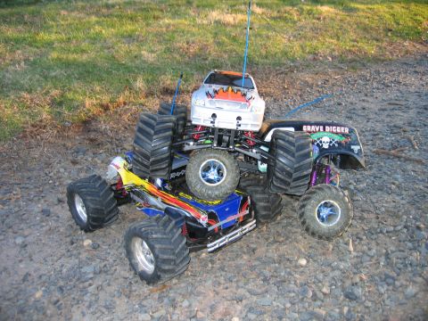 1996 Ford Crown Victoria - A pair of RC monster trucks, one labeled 'Grave Digger', are entangled in a playful collision on a dirt and gravel ground.