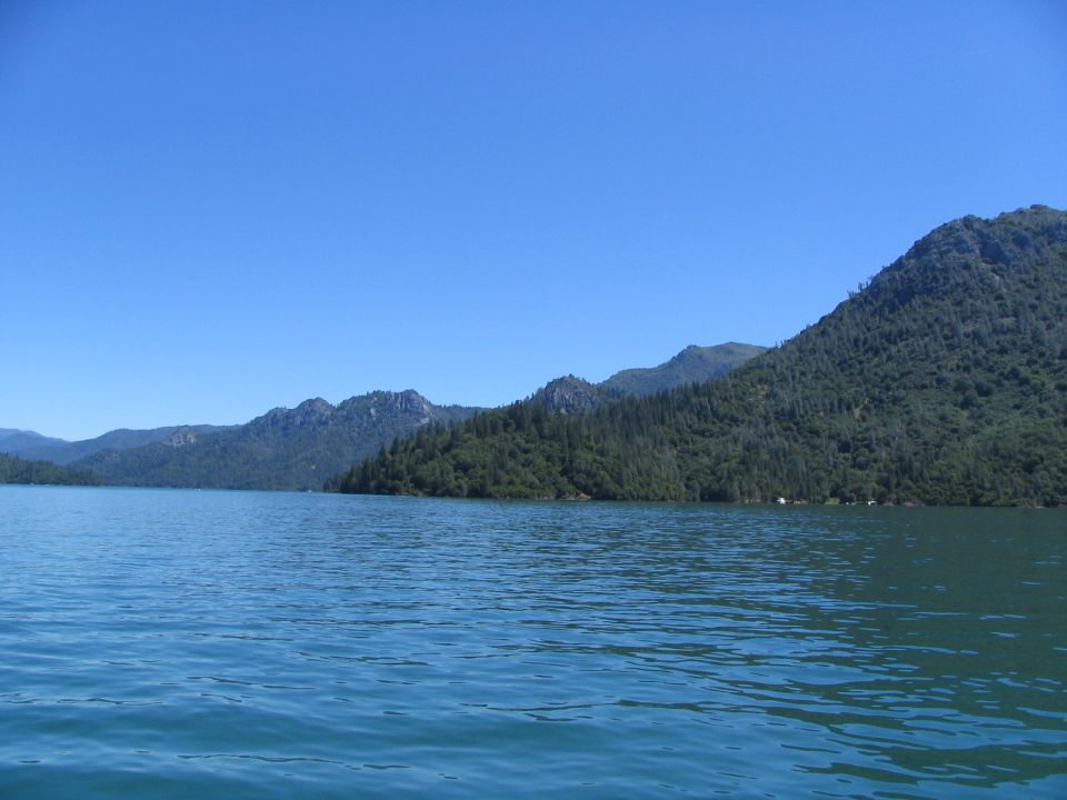 Blue lake water with tree-covered mountains under a clear blue sky. Supermotors.net logo in lower right