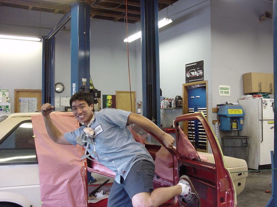 Guy posing in a garage next to a car being worked on. Another car is partially visible on the left