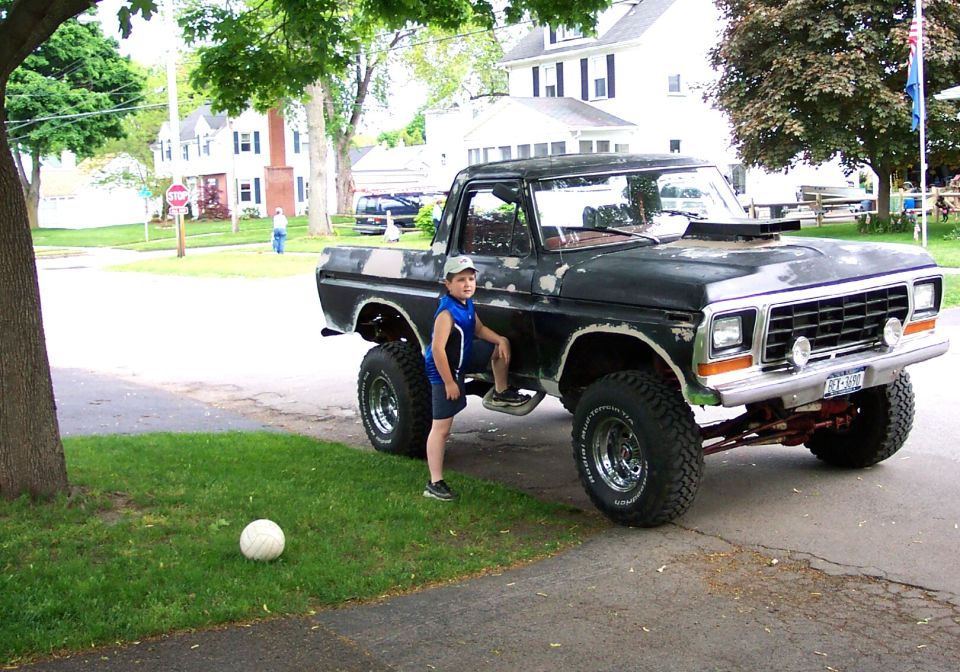 Boy in blue jersey beside a tall lifted black Ford Bronco with a soccer ball in the yard