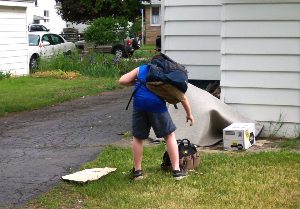 Boy with backpack bending over a tool bag beside a house with a generator and rolled carpet