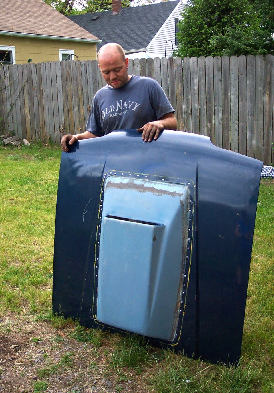 Man in gray Old Navy shirt holding a dark car hood with a riveted cowl scoop in a backyard