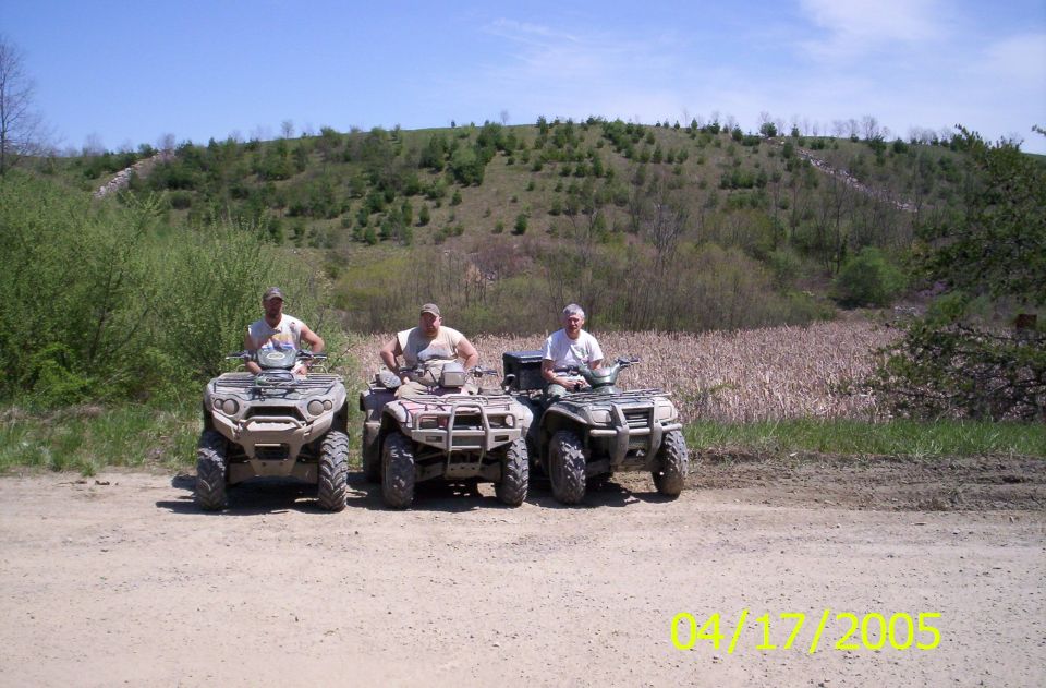 Three men on ATVs parked side by side on dirt with a green hillside behind