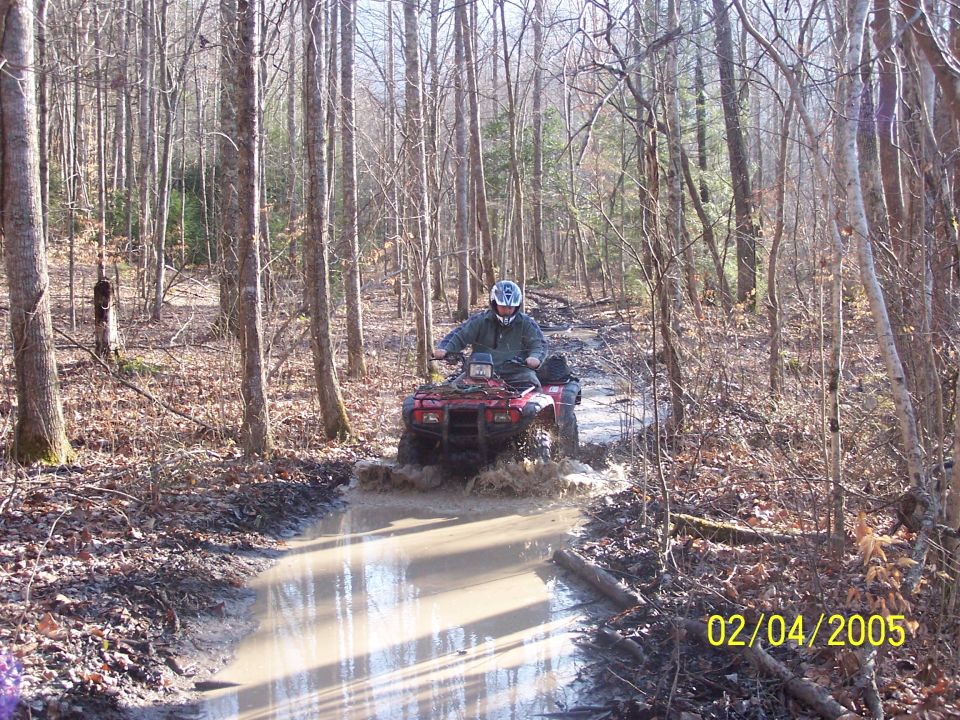 Rider on red utility ATV crossing a muddy puddle on a wooded trail, dated 02/04/2005