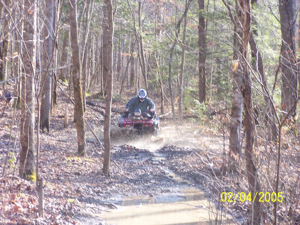 Rider on a red ATV splashing through a muddy trail puddle in bare winter woods