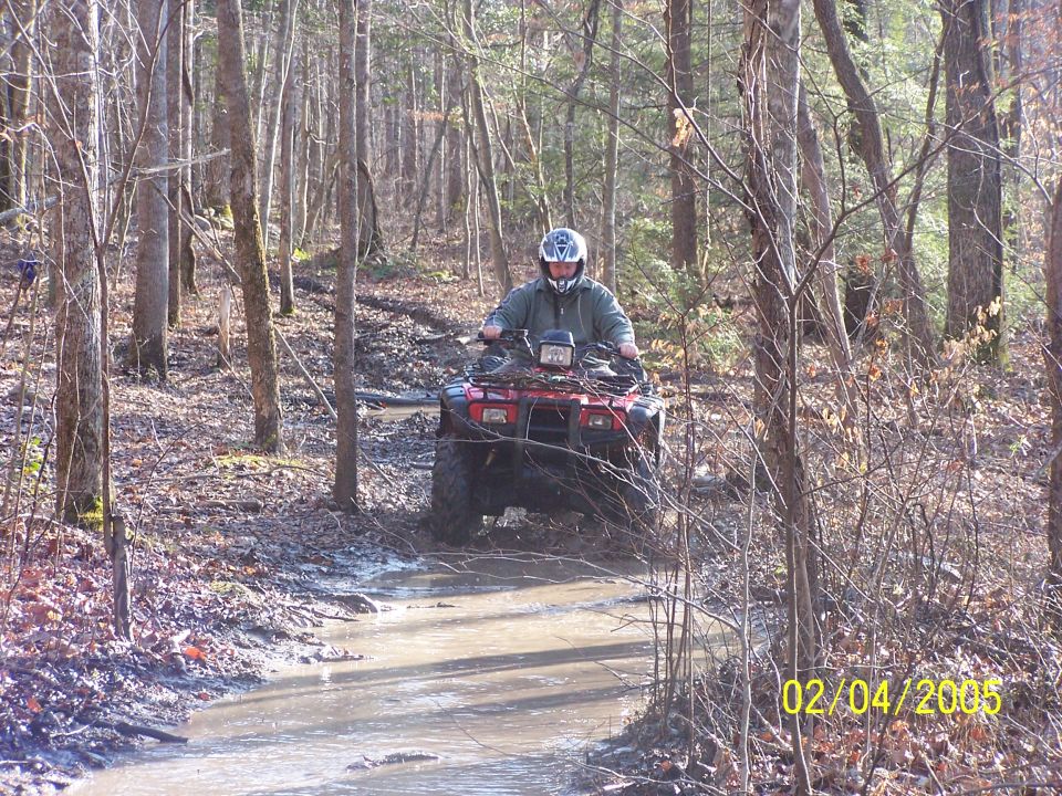 Red ATV with rider crossing a muddy forest trail with water in the ruts.