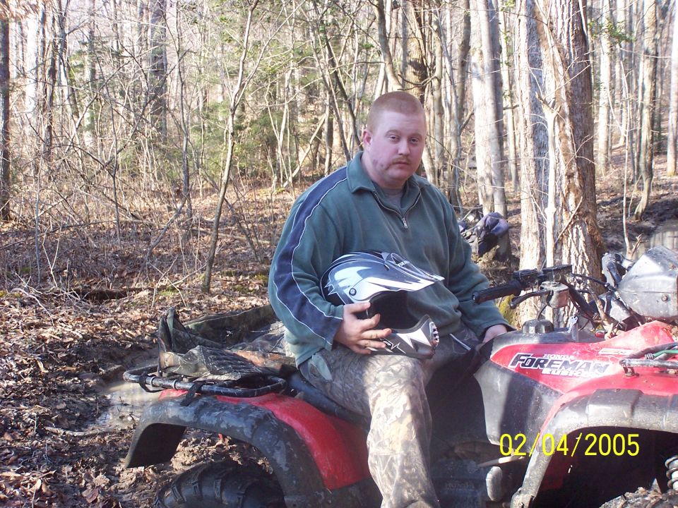 Man holding helmet sits on red Honda Foreman ATV in muddy woods