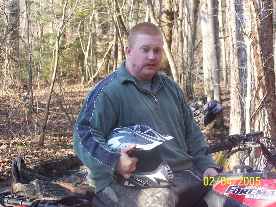 Man holding a Fulmer helmet while seated on a red Honda Foreman ATV in the woods.