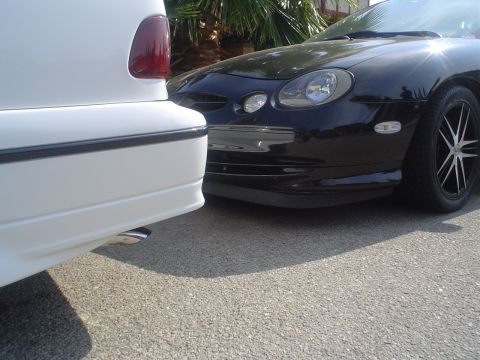 1993 Ford Taurus - The front of a black car is visible next to a white car, parked on asphalt.