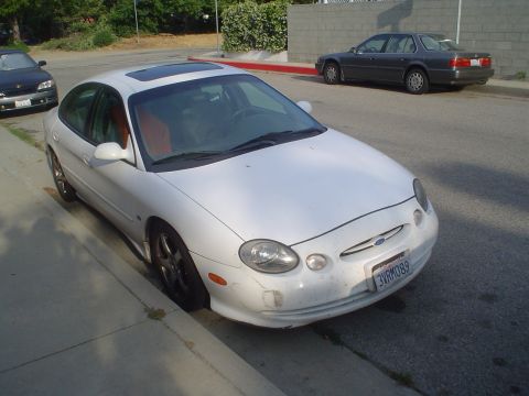 1993 Ford Taurus - A white Ford Taurus is parked curbside, with a dark sedan visible in the background.