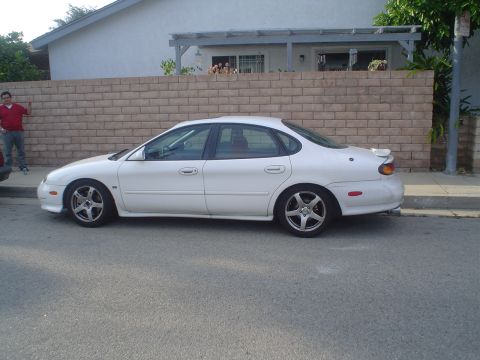1993 Ford Taurus - A white sedan is parked on the side of the road, with a person standing nearby.